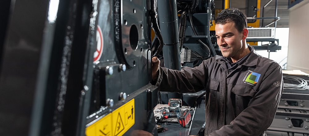 „Servicemonteur voert onderhoudswerkzaamheden uit aan een grote bouwmachine in een werkplaatsomgeving.