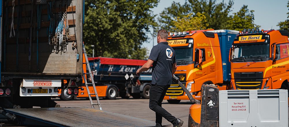 Een medewerker verplaatst materiaal naast een open vrachtwagenoplegger op het terrein van transportbedrijf Ter Horst, met meerdere oranje vrachtwagens op de achtergrond.