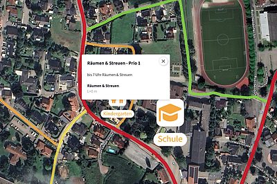 Luftbildkarte eines Wohngebiets mit farblich markierten Straßen für Räum- und Streueinsätze. Ein Hinweisfenster zeigt ‚Räumen & Streuen – Prio 1‘ sowie Zeitangaben; Symbole markieren Orte wie Kindergarten und Schule