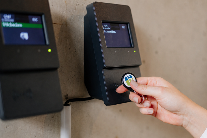 Eine Person hält einen kleinen Chip an ein digitales Zeiterfassungsterminal an der Wand. Auf dem Display ist eine Anmeldefunktion zu sehen; daneben befindet sich ein weiteres Terminal.